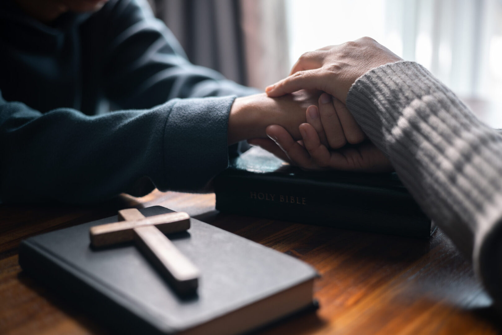 Two people holding hands over a Bible with a wooden cross beside it.