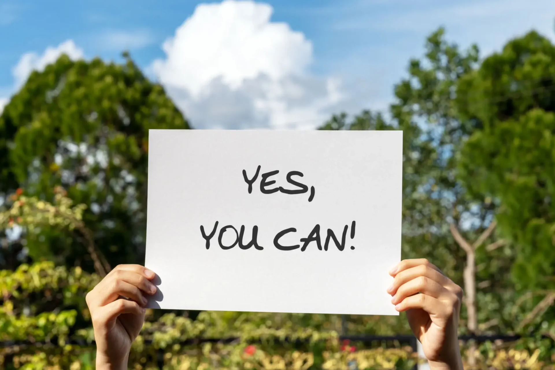 A person holding a motivational sign that says, 'YES, YOU CAN!' outdoors.